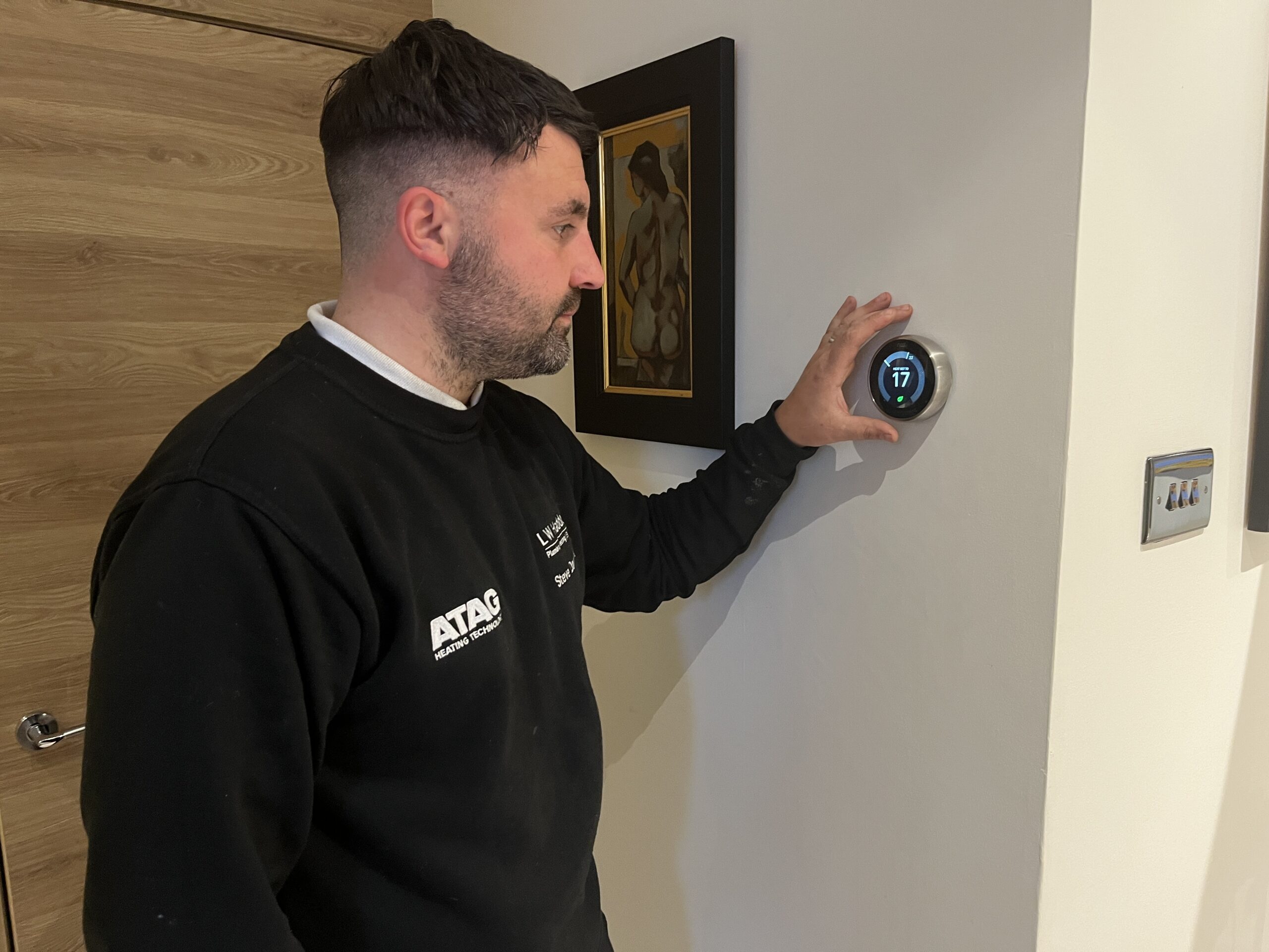 An engineer from LW. Haddow Plumbing and Heating adjusts a smart thermostat on a wall after a smart heating installation.