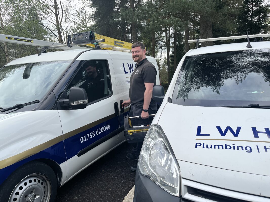 An engineer from L.W. Haddow Plumbing and Heating is standing next to company vans with a tool bag.