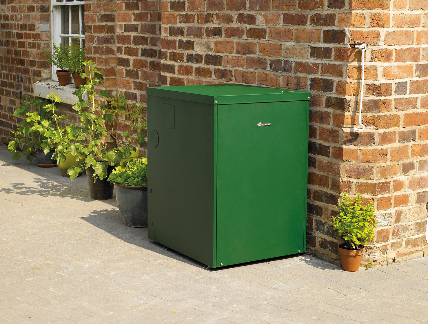 outdoor oil boiler in green casing installed against a traditional red brick house, surrounded by potted garden plants